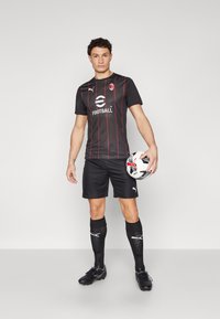 Čierny futbalový dres s červenými pruhmi, logo na hrudi, v kombinácii s čiernymi šortkami, ponožkami a kopačkami, držiaci čierno-biely futbal.