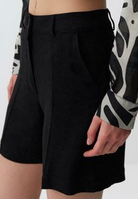 Pantaloni corti neri in lino con pieghe, dotati di tasche laterali diagonali e parte anteriore piatta. Una camicia a maniche lunghe con un motivo è parzialmente visibile.