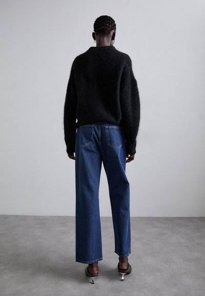 Zwart, fluffy trui met wijde mouwen, gecombineerd met hooggesneden, recht gesneden blauwe jeans. Model draagt bruine hakschoenen, gezien vanuit de achterkant.
