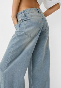 Kvinna i högmidjade, ljusblå vida jeans med händerna i fickorna, sedd bakifrån från midjan och ner mot en enkel bakgrund.