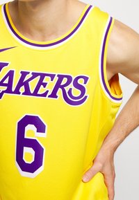 Žlutý basketbalový dres nese fialový nápis "LAKERS" a fialové číslo 6. Vyroben z prodyšné síťoviny s kulatým výstřihem a průramky.