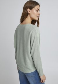 Helles grünes, oversized Strickpullover mit einer weichen Textur, weitem Ausschnitt und überschnittenen Schultern, kombiniert mit blauen Jeans.