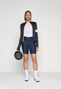 Cycling outfit featuring a navy jersey with pink trim, a white mesh insert, and navy shorts. Model holds a black helmet and wears white socks.