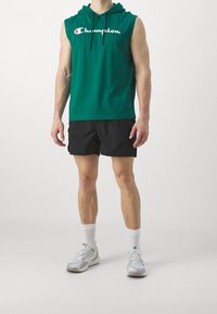 Zelená bezrukávová mikina z bavlny s bílým logem na přední straně. V kombinaci s černými šortkami a světle šedými sportovními botami.