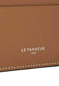 Materiale in pelle marrone con una superficie testurizzata; presenta cuciture bianche e un logo in rilievo argentato "LE TANNEUR 1898."