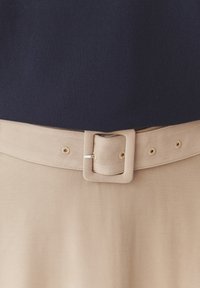 Marineblauwe trui met een beige stoffen riem, voorzien van een vierkante metalen gesp en knoopsgaten, tegen een gladde textielachtergrond.