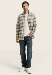 Geruite flanellen shirt in beige en bruine tinten, gecombineerd met een witte T-shirt en blauwe denim jeans. Witte sneakers maken de outfit af.