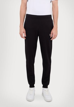 Homme portant un pantalon de jogging noir avec des poignets et une taille élastiques, des baskets blanches et une chemise blanche, debout devant un fond uni.