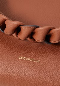 Smeđa kožna torba s pletenim dizajnom duž vrha. Ima zlatni logo "Coccinelle" i teksturiranu površinu.
