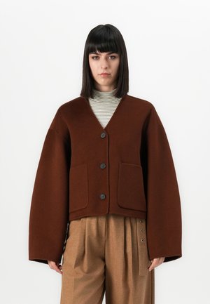 Žena s ravnom crnom kosom, koja nosi smeđu oversized jaknu s dugmadi, prugasti rolak i bež hlače visokog struka.