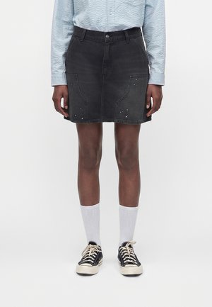 Person iført en sort denim mininederdel med lommer, en lyseblå langærmet skjorte, hvide sokker og sorte lavtop-sneakers.