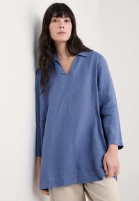 Blauwe linnen tuniek met een V-hals en lange mouwen, met een losse pasvorm en zijsplitten aan de zoom, tegen een effen achtergrond.