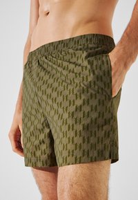 Shorts da bagno color khaki con motivo geometrico su tutta la superficie, vita elastica e tasche laterali, realizzati in tessuto leggero e testurizzato.