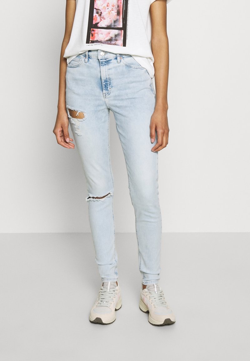 Jeans skinny de color azul claro con detalles de rasgados en las rodillas, combinados con una camiseta blanca con un gráfico floral rosa, y zapatillas ligeras.