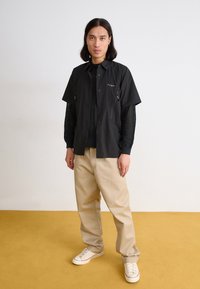 Black short-sleeve shirt with front buttons and pockets, layered over a black mesh top, paired with light tan wide-leg pants and white sneakers.
