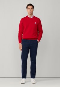Sudadera roja con logo blanco, llevada sobre una camisa de cuadro claro con cuello. Combinada con pantalones azul marino y zapatillas blancas. Fondo liso.