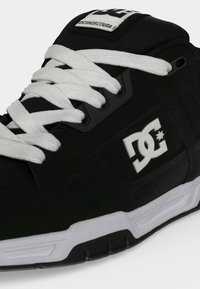 Κοντινή όψη μαύρου και άσπρου sneaker της μάρκας DC με άσπρα κορδόνια και λογότυπο στο πλάι και στη γλώσσα.