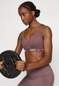 Καφέ αθλητικό σουτιέν με φαρδιές τιράντες και λευκή λωρίδα με το λογότυπο της Nike, σε συνδυασμό με κολάν ψηλόμεσο. Το άτομο κρατά μια μαύρη δισκοειδή βαράκι.