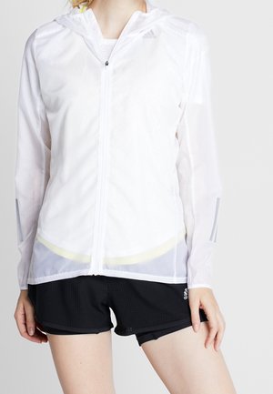 Vrouw die een witte, lichte Adidas jas met rits en capuchon draagt, en zwarte Adidas sportshorts tegen een effen achtergrond.
