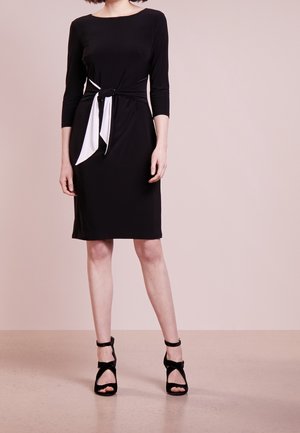 Femme portant une robe noire mi-longue aux manches trois-quarts avec un détail de nœud blanc à la taille, assortie de sandales à talons hauts noires.