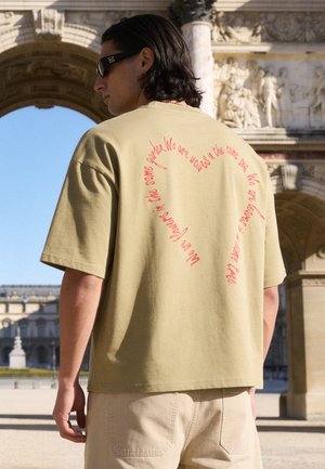 Homme aux cheveux foncés et lunettes de soleil portant un t-shirt beige oversize avec un texte rouge formant un cœur dans le dos, debout à l'extérieur près d'une arche historique.