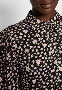 Blusa com estampa floral em fundo preto, pequenas flores cor-de-rosa com folhas verdes, colarinho pontudo e detalhes de botões na frente.