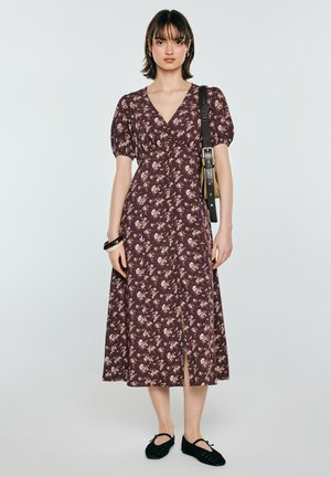 Kobieta w midi sukience w kwiaty z bufkami, czarnych balerinach, czarnej torebce na ramię oraz z nawarstwionymi bransoletkami na nadgarstku.