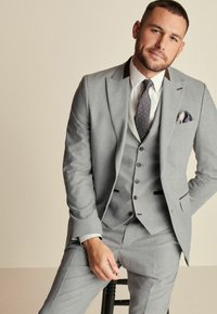Traje gris de tres piezas con un corte ajustado, que presenta una tela texturizada, solapas de muesca, botones oscuros y una corbata de estampado. Acompañado de un pañuelo de bolsillo.