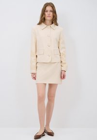 Veste beige ajustée avec fermeture à boutons et broderie florale sur les manches, associée à une jupe assortie et des chaussures à enfiler marron.