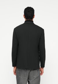 Blazer preto com um corte slim, lapelas recortadas e dois botões. Abertura nas costas para movimento, feito de um tecido suave, combinado com jeans cinzentos.