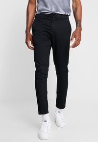 Chino neri su misura realizzati in un tessuto liscio, caratterizzati da una vestibilità slim e passanti per la cintura, abbinati a sneaker bianche e una camicia a righe.