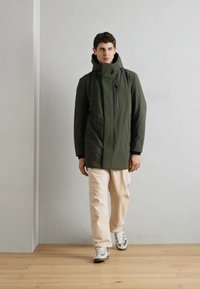 Donkergroene waterdichte jas met hoge kraag, voorkant met rits en meerdere zakken, gedragen over lichte beige broek en grijze sneakers.