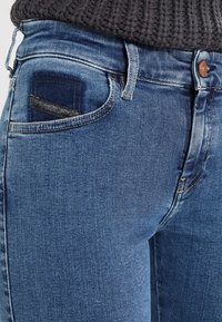 Närbild på blå denimjeans med en synlig myntficka, kopparknapp och en mörk texturerad tröja instoppad i midjan.