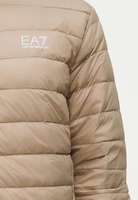 Bézs színű steppelt puffertélkabát vízszintes csíkokkal. A bal felső részen fehér EA7 logó található. Simogató, szintetikus anyagú felület.