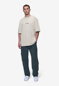 Beżowy, oversize t-shirt z napisem "PEGADOR", połączony z ciemnozielonymi spodniami cargo z wieloma kieszeniami i białymi trampkami.