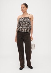 Zwart en beige panterprint top met dunne bandjes, gecombineerd met donkerbruine rechte broek en zwarte flats; houdt een witte tas vast.