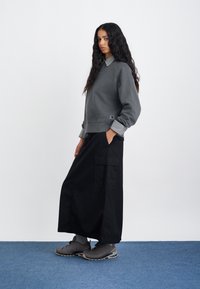 Šedý mikina s rebrovaným výstrihom a pruhovanými manžetami, spárovaná s dlhou čiernou sukňou s vreckami a šedými teniskami. Jednoduché pozadie.