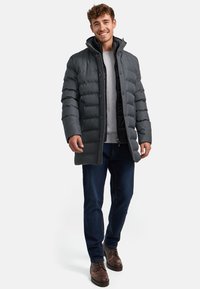 Donkere grijze pufferjack met horizontaal gestikt ontwerp, hoge kraag, knoopsluiting en voorzakken. Draagt over een lichtgrijze top en jeans.