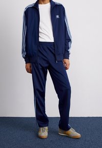 Chándal navy con rayas azul claro, que incluye una chaqueta con cremallera y pantalones con cintura elástica. El conjunto se combina con zapatillas grises con acentos amarillos.