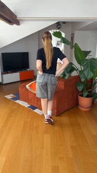 Γυναίκα με μακριά αλογοουρά στέκεται με τα χέρια στη μέση, βλέποντας έναν κόκκινο καναπέ· μεγάλο πράσινο φυτό δίπλα του και τηλεόραση στο βάθος.