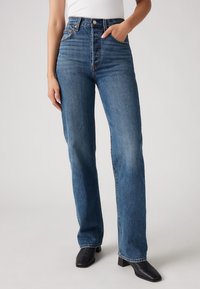 Kvinna klädd i högmidjade raka blå jeans och svarta klackstövlar med fyrkantig tå, med ena handen i fickan och en vit topp.