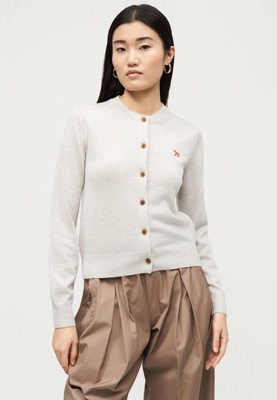 Νέα γυναίκα με μπεζ ζακέτα με ξύλινα κουμπιά και καφέ πλισέ παντελόνι, στέκεται μπροστά σε απλό φωτεινό φόντο.