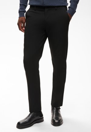 Homme portant un pantalon noir slim, des bottines Chelsea en cuir noir et un pull en tricot bleu, debout les mains dans les poches.