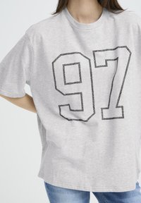 Pelēka īsroku krekla T-krekls ar lielu melnu rhinestone grafiku, kurā ir skaitlis "97" biezā, ārpusē zīmētā dizainā.