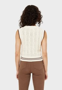 Vrouw met krullend bruin haar, gekleed in een mouwloos crèmekleurig kabelbreisel truitje en bruine broek, met haar rug naar de camera gekeerd, tegen een effen achtergrond.