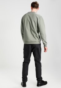 Felpa grigia con collo rotondo e polsini a costine, abbinata a pantaloni scuri e scarpe nere, vista da dietro su uno sfondo neutro.