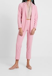 Femme portant un costume rose ajusté avec un pantalon court, un crop top beige côtelé, des boucles d'oreilles turquoise et des chaussures plates nude, debout contre un mur blanc.