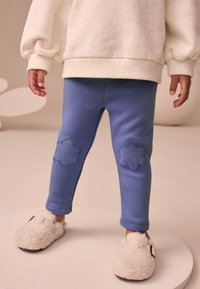 Colanți albaștri din bumbac cu pete texturate în formă de flori pe genunchi, asortați cu papuci bej pufoși. Ținuta prezintă un design relaxat și casual.