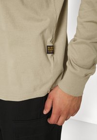 Camicia beige a maniche lunghe con etichetta G-Star Raw nera e gialla vicino all'orlo, indossata sopra pantaloni neri con tasca, con la mano appoggiata di lato.