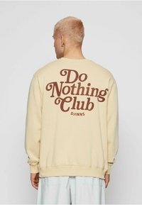 Sudadera color crema con un ajuste relajado, que presenta una gran impresión de texto en estilo retro de color marrón que dice "Club de No Hacer Nada" en la parte posterior.